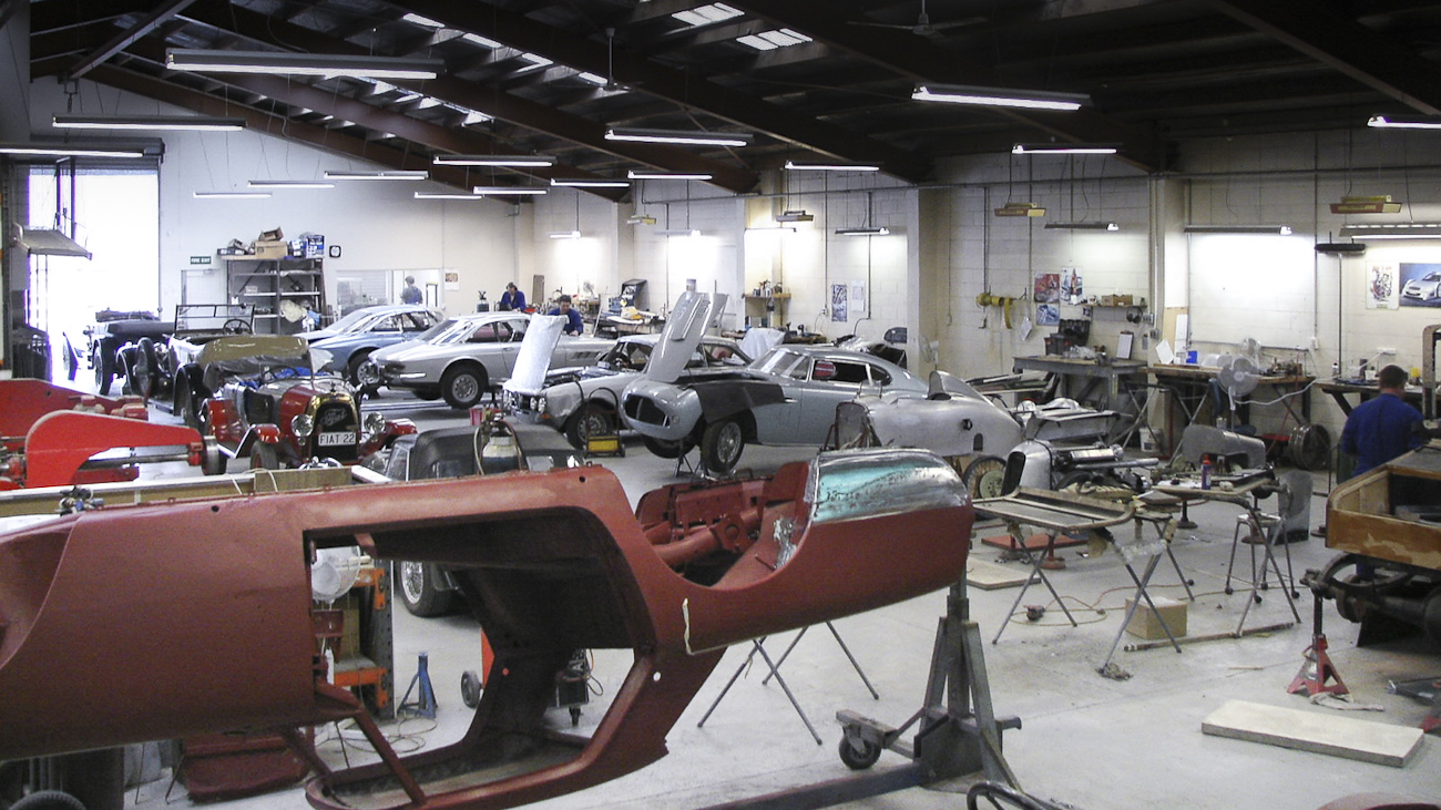 Wide view of in-progress restorations of rare classics in the Panel Shop and at Auto Restorations. Includes a Siata 280CS Balbo, an Alfa Romeo, a Fiat, a pair of Ferrari in the background and a Ferrari Lusso suspended upside down in the cradle in the foreground.