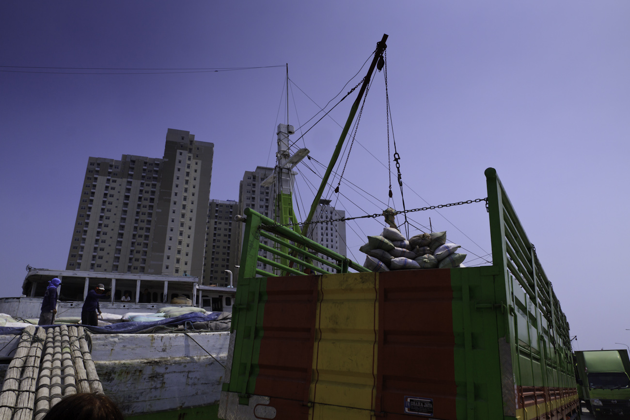 A deck crane aboard a lamba type of motorised Pinisi offloading a pallet of palm kernel cake cattle feed from Borneo to a waiting freight truck at the port of Sunda Kelapa, old harbour of Jakarta.