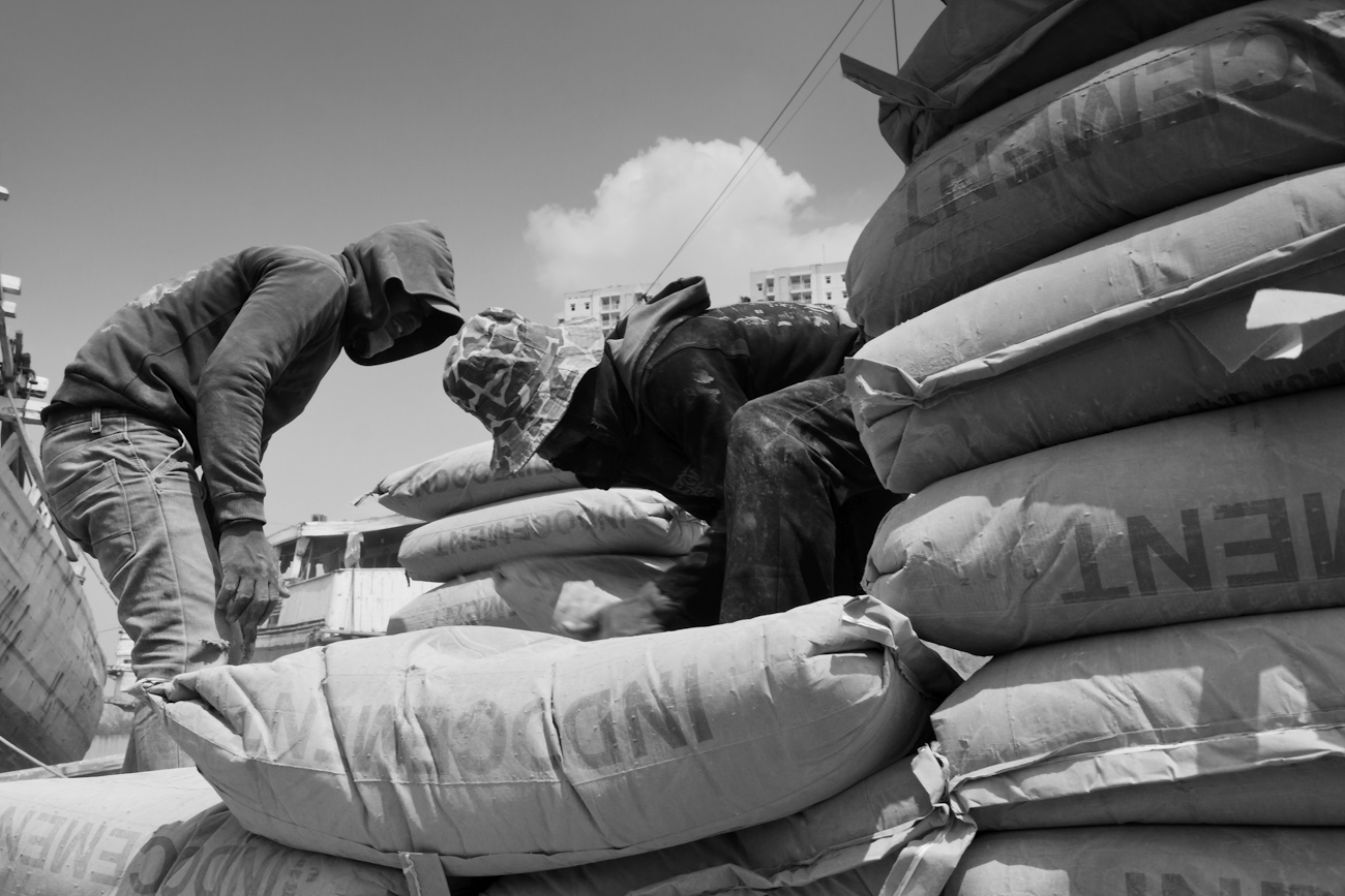 Coolies on the flat deck of a commercial truck alongside a Pinisi freighter load 50 kg bags of Portland Cement onto a wooden pallet to be lifted by crane into the freighter.