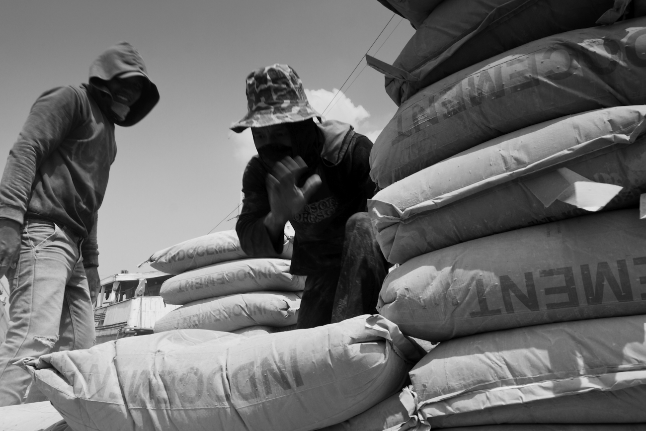 Coolies on the flat deck of a commercial truck alongside a Pinisi freighter load 50 kg bags of Portland Cement onto a wooden pallet to be lifted by crane into the freighter.