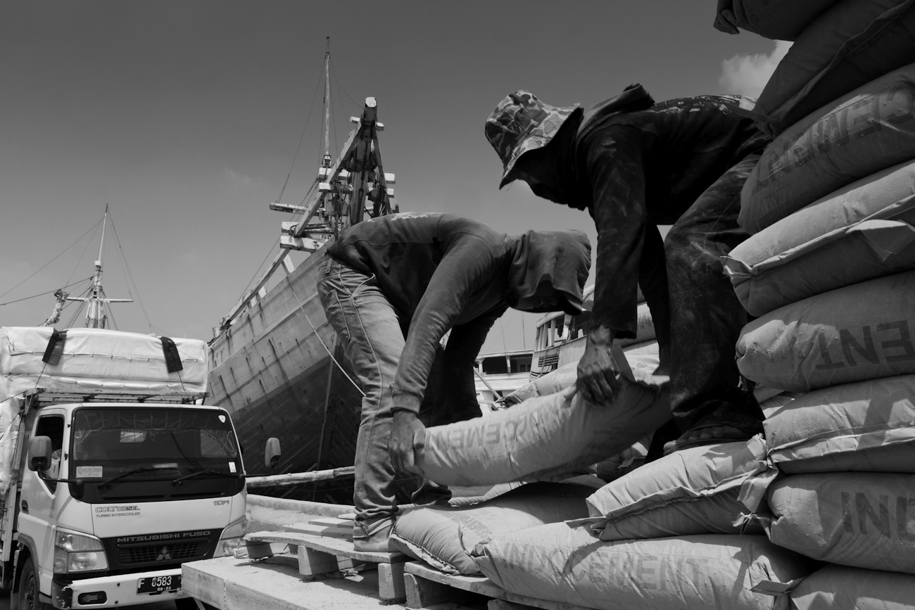 Coolies on the flat deck of a commercial truck alongside a Pinisi freighter load 50 kg bags of Portland Cement onto a wooden pallet to be lifted by crane into the freighter.