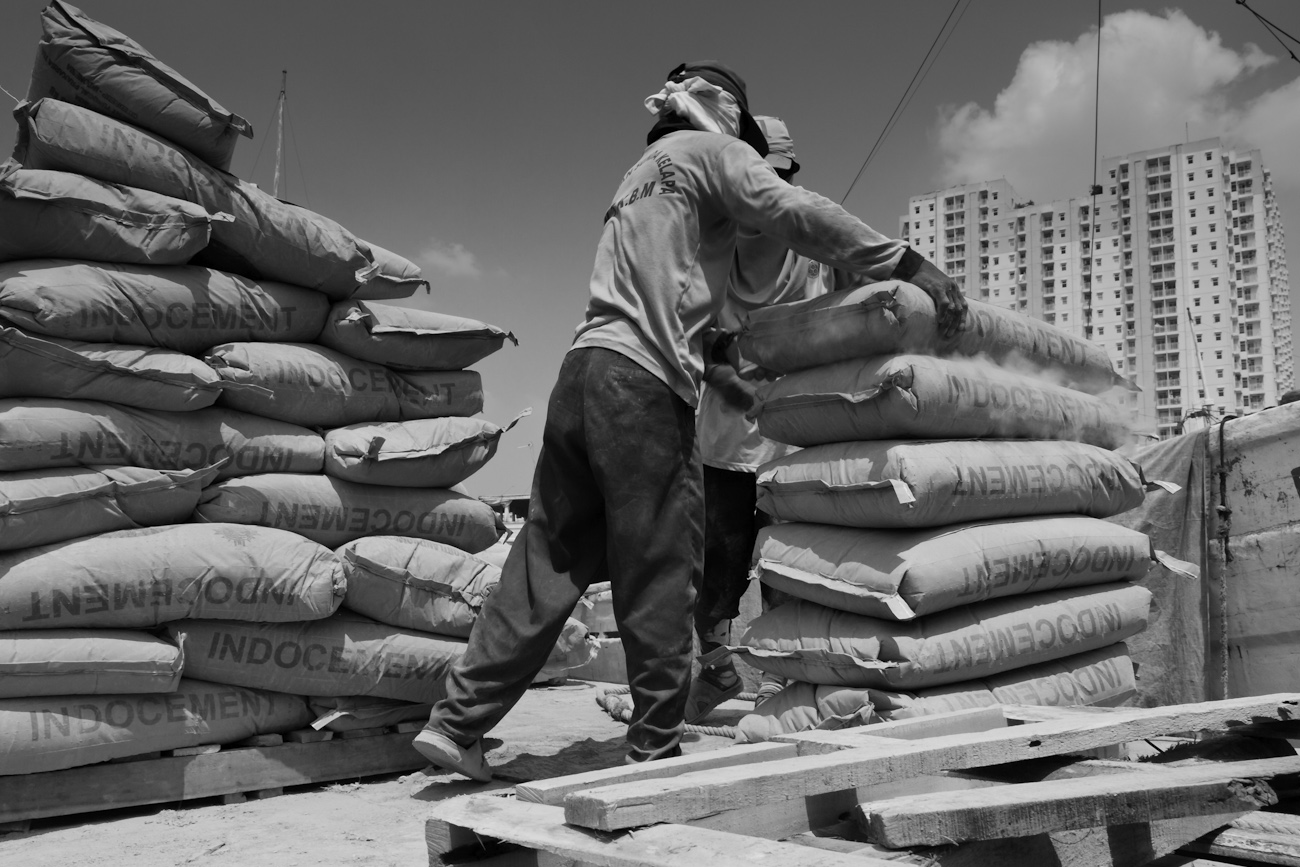 Coolies on the flat deck of a commercial truck alongside a Pinisi freighter load 50 kg bags of Portland Cement onto a wooden pallet to be lifted by crane into the freighter.