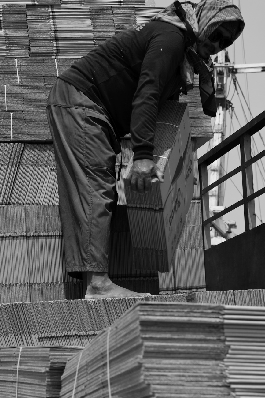 Coolie on the flat deck of a commercial truck alongside a lamba Pinisi freighter loads flat-packed cardboard boxes onto a wooden pallet to be lifted by crane onto the freighter.