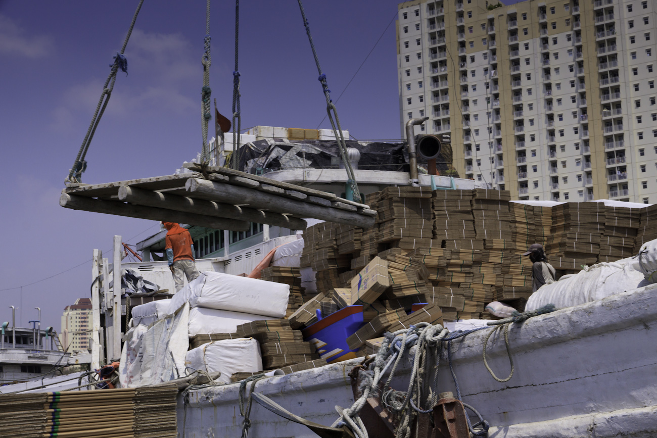 Pinisi freighter deck crane cargo operations, empty crane lifting platform returns to deck of freight truck for the hand loading of another load of cardboard flat-packed boxes.