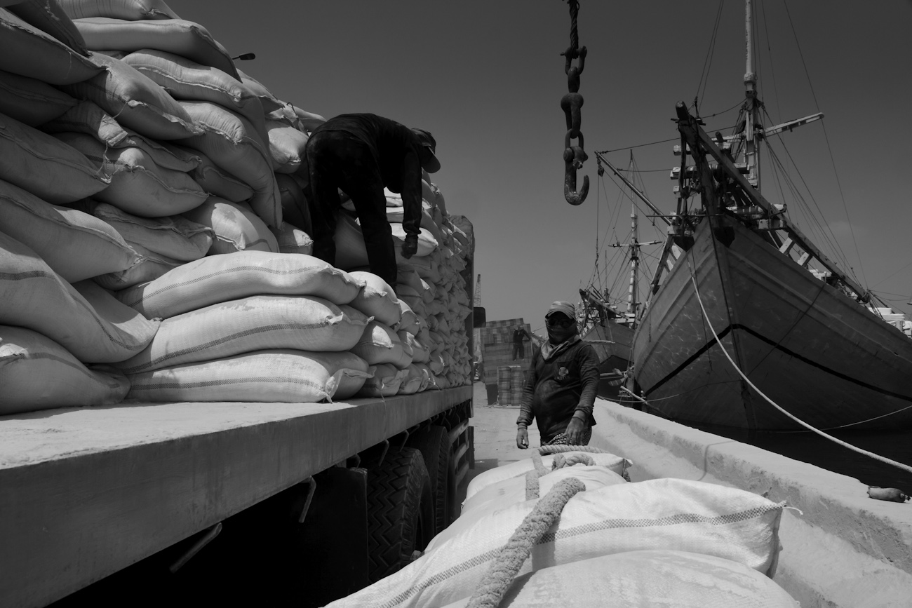 After the deck crane aboard a motorised Pinisi offloads a pallet of palm kernel cake cattle feed from Borneo, the 20 kg bags are hand stacked by a pair of coolies onto the trailer of a waiting freight truck. Port of Sunda Kelapa, old harbour of Jakarta, Indonesia.