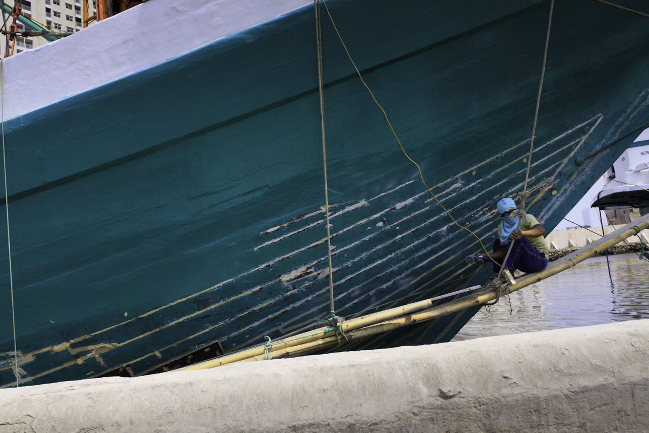 The prow of a motorised Pinisi is being sanded, scraped back and cracks filled with epoxy by a coolie prior to repainting with roller and brush applied lacquer. Sunda Kelapa, old harbour, Jakarta, Indonesia.