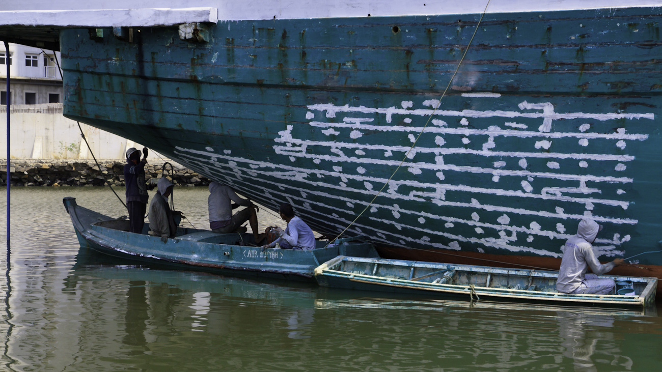 The stern of a motorised Pinisi is being sanded, scraped back and cracks filled with epoxy and repainted with roller and brush applied lacquer above the waterline by 5 coolies working off two dinghis.