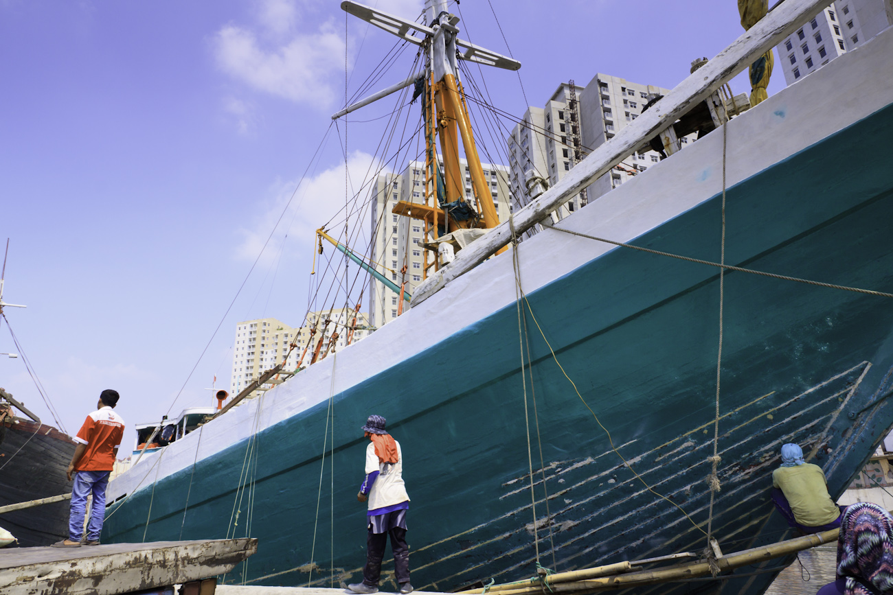 The prow of a motorised Pinisi is being sanded, scraped back and cracks filled with epoxy prior to being repainted with roller and brush applied lacquer above the waterline by a coolie working off a suspended plank Sunda Kelapa, old harbour, Jakarta, Indonesia.