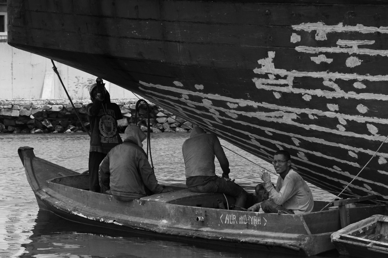 The stern of a motorised Pinisi is being sanded, scraped back and cracks filled with epoxy above the waterline by 5 coolies working off two dinghis. Sunda Kelapa, old harbour, Jakarta, Indonesia.