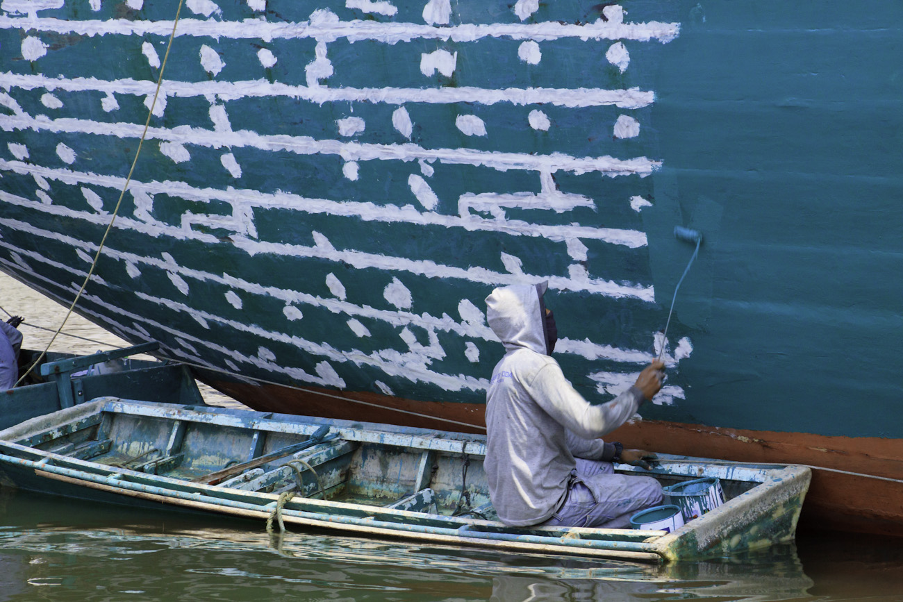 The stern of a motorised Pinisi being repainted with roller and brush applied lacquer above the waterline by a coolie working off a dinghi.