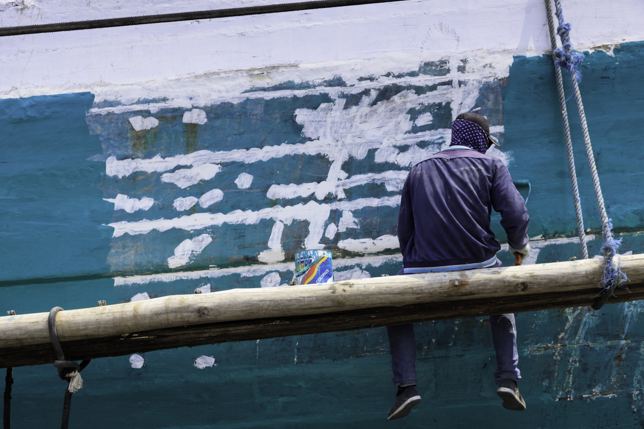 The midships of a motorised Pinisi being repainted with roller and brush applied lacquer above the waterline by a coolie working off a suspended plank