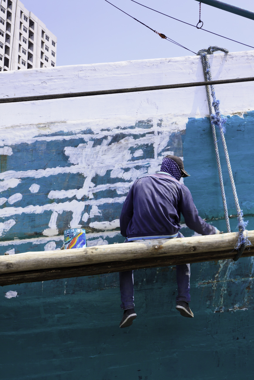 The midships of a motorised Pinisi being repainted with roller and brush applied lacquer above the waterline by a coolie working off a suspended plank.