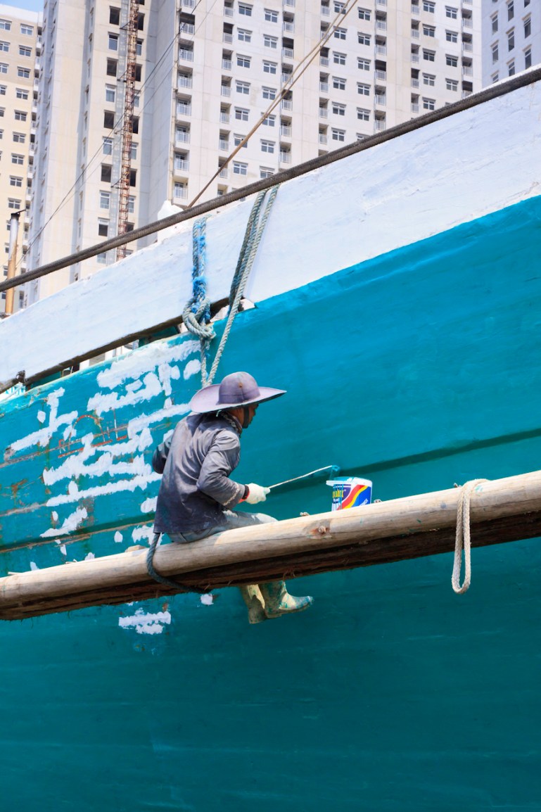 The midships of a motorised Pinisi being repainted with roller and brush applied lacquer above the waterline by a coolie working off a suspended plank.