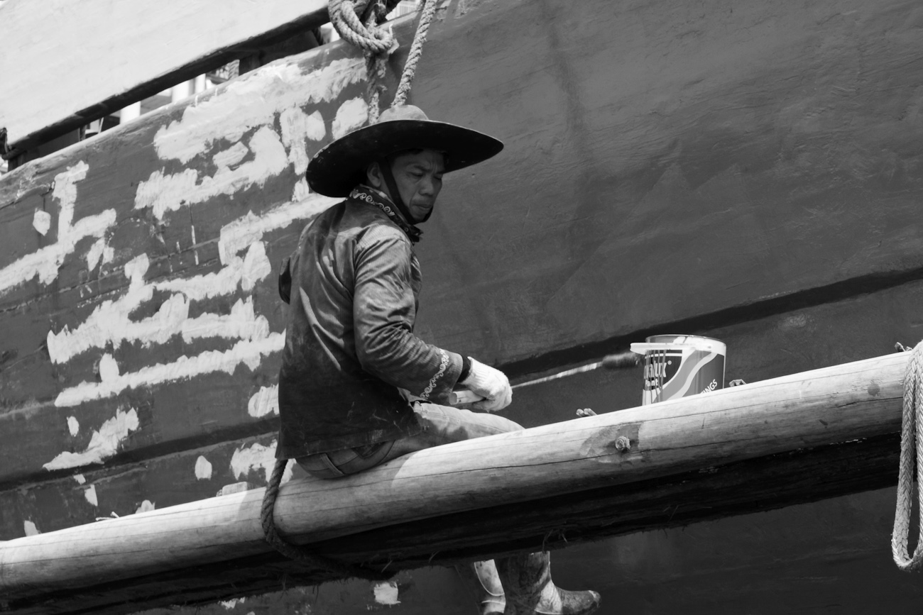 The midships of a motorised Pinisi being repainted with roller and brush applied lacquer above the waterline by a coolie working off a suspended plank.
