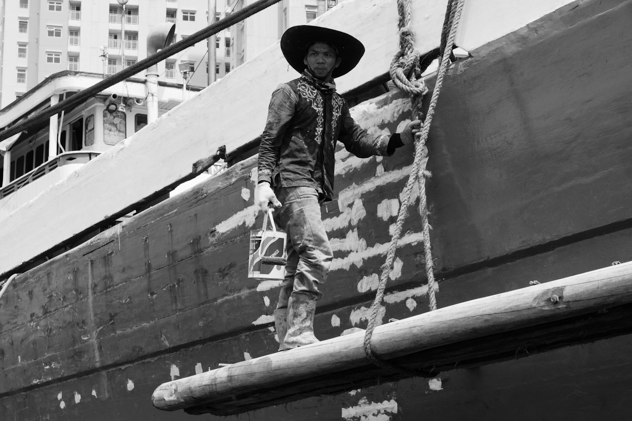 The midships of a motorised Pinisi being repainted with roller and brush applied lacquer above the waterline by a coolie working off a suspended plank.