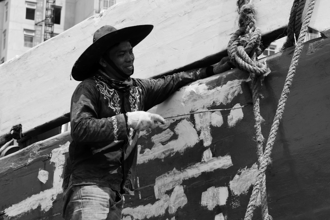 The midships of a motorised Pinisi being repainted with roller and brush applied lacquer above the waterline by a coolie working off a suspended plank.