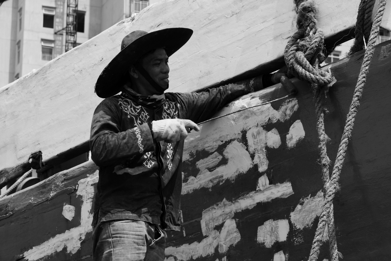 The midships of a motorised Pinisi being repainted with roller and brush applied lacquer above the waterline by a coolie working off a suspended plank.