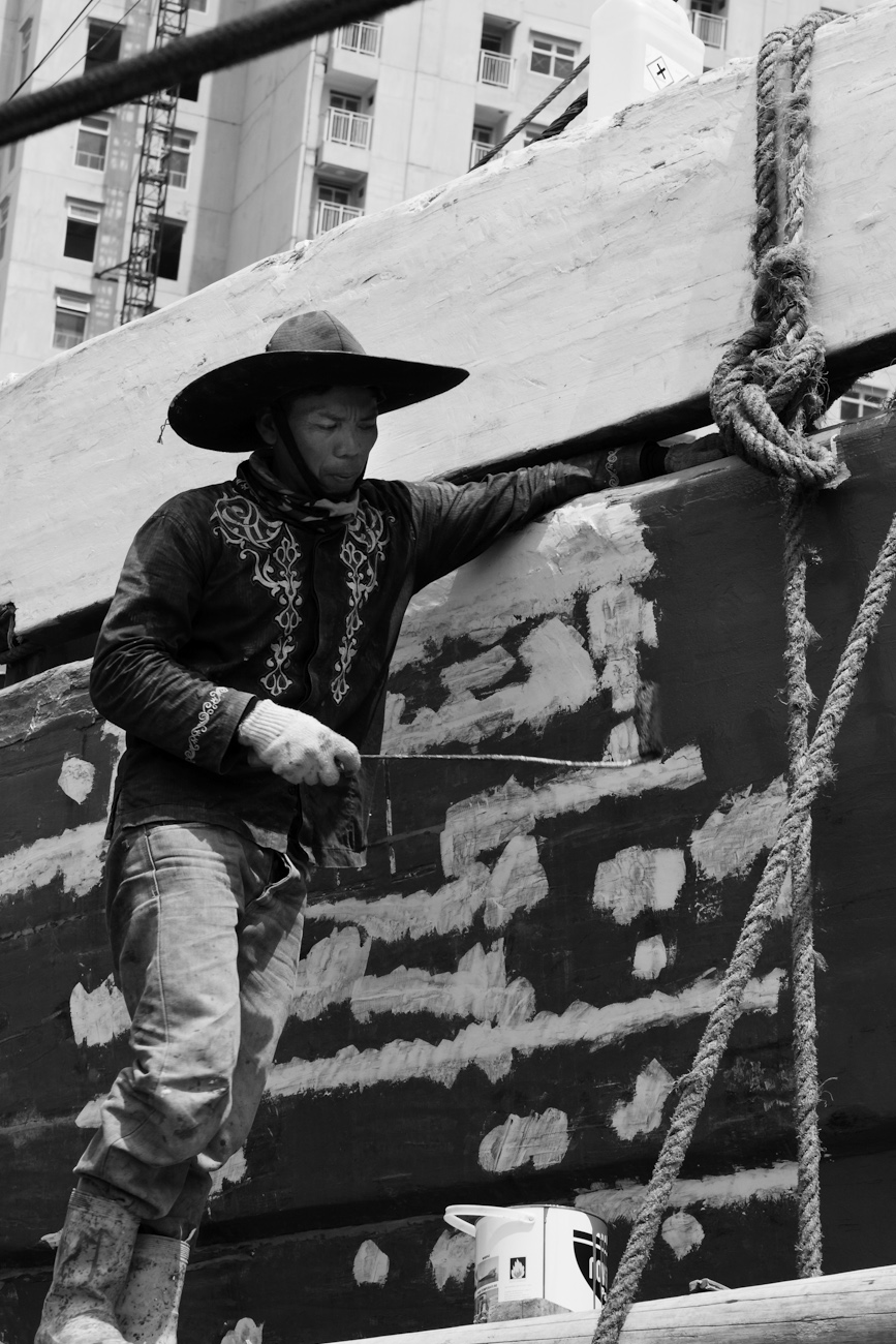 The midships of a motorised Pinisi being repainted with roller and brush applied lacquer above the waterline by a coolie working off a suspended plank.