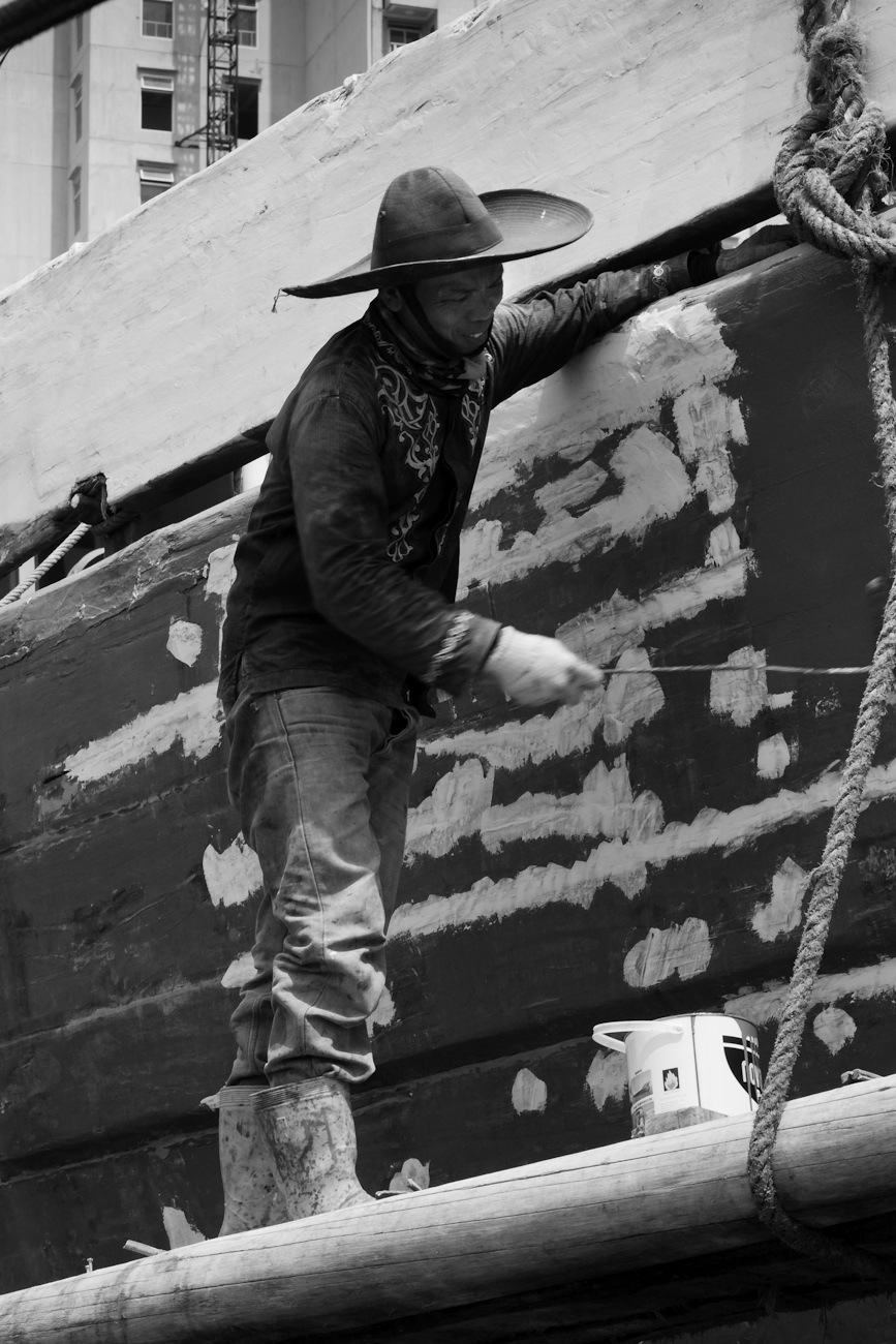 The midships of a motorised Pinisi being repainted with roller and brush applied lacquer above the waterline by a coolie working off a suspended plank.