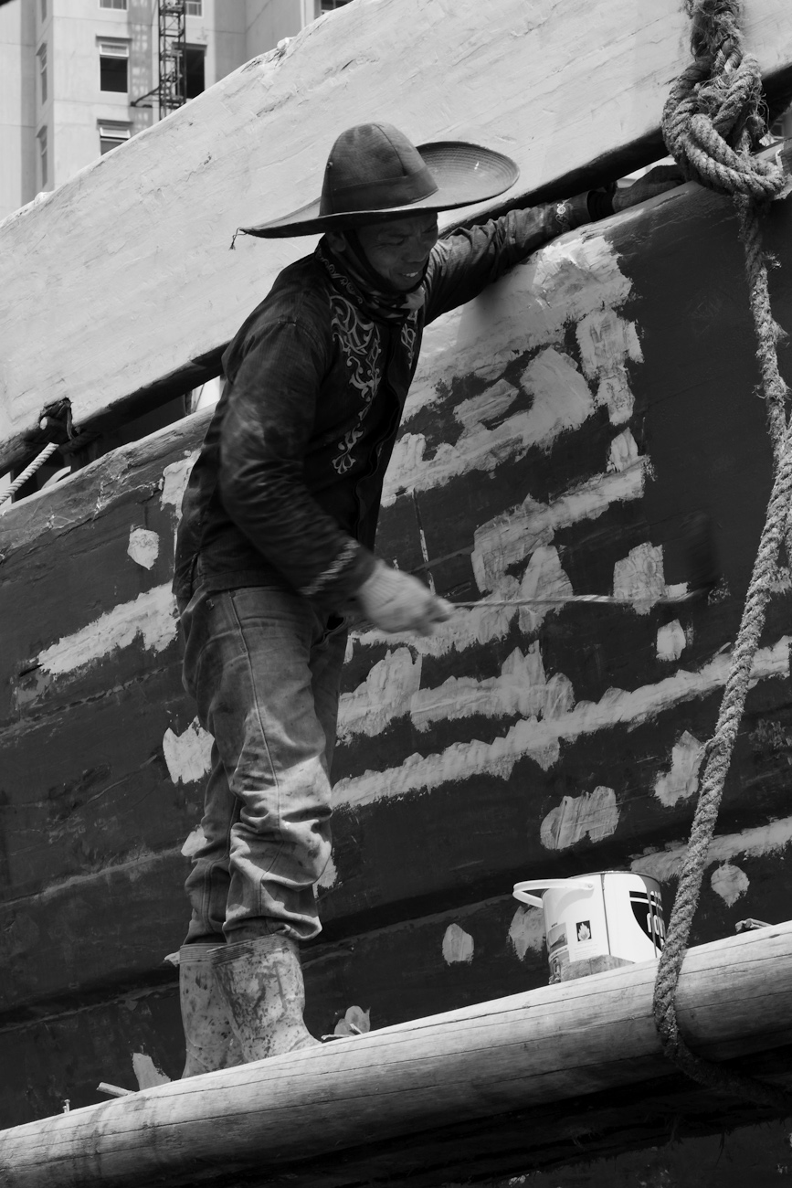 The midships of a motorised Pinisi being repainted with roller and brush applied lacquer above the waterline by a coolie working off a suspended plank.