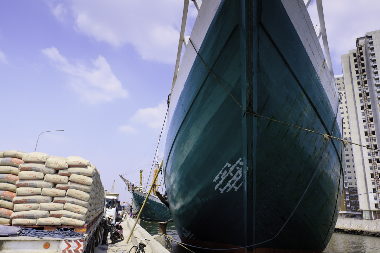 Lamba type of motorised Pinisi line up alongside the wharf at Sunda Kelapa, old harbour of Jakarta. Freight trucks laden with 50 kg bags of Portland cement cargo await loading by a combination of deck crane and hand stacking by coolies.