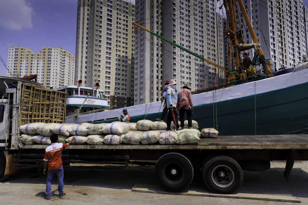 20 kg sacks of cargo being offloaded by deck crane and hand-stacked by coolies on the flat deck of a waiting freight truck.