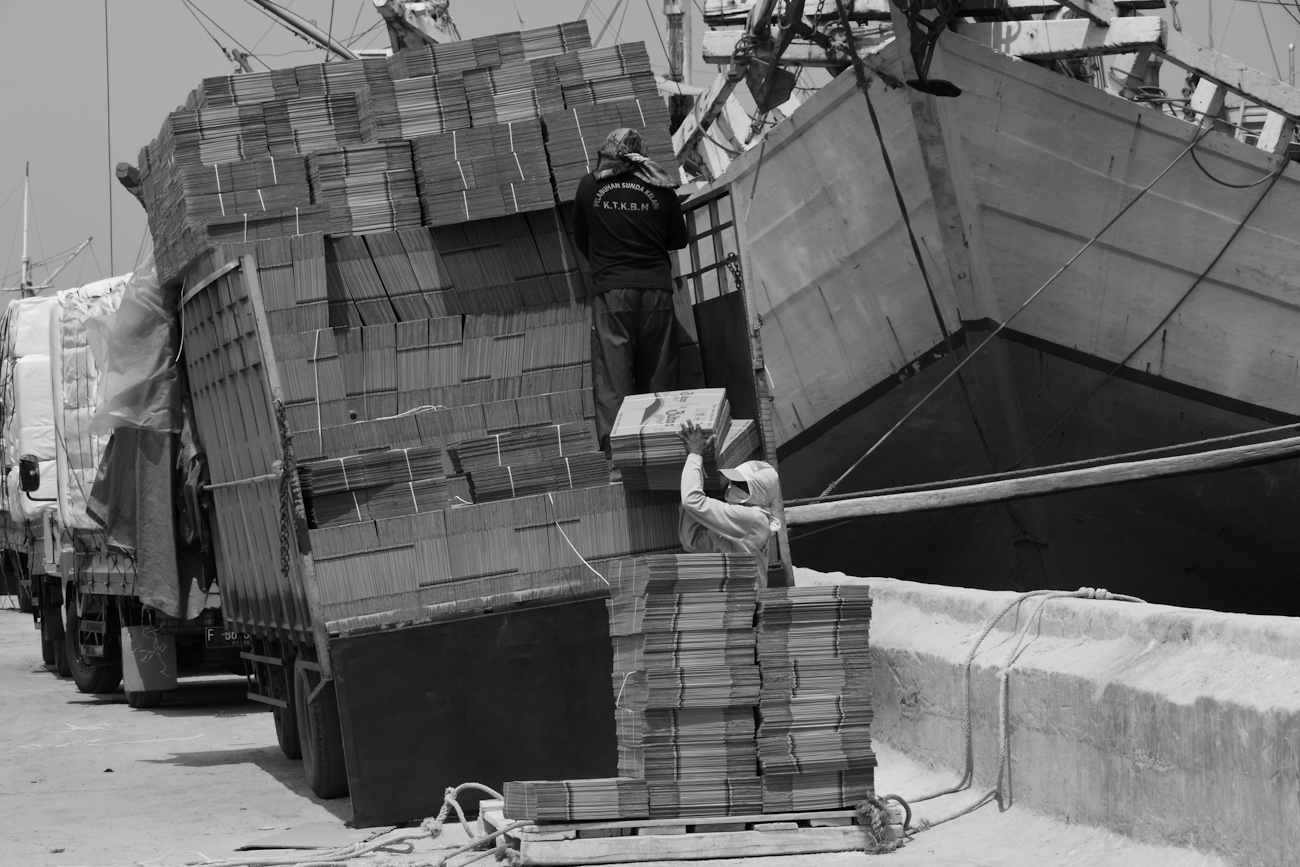 A coolie hand loads flat-packed cardboard boxes onto a wooden pallet on the wharf prior to loading into an awaiting Pinisi freighter using a deck crane.