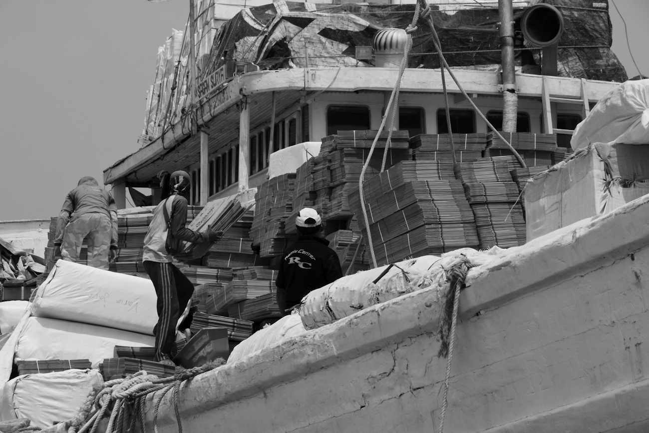 A pallet load of flat-packed cardboard boxes is loaded onto the deck of an awaiting Pinisi freighter using a deck crane.
