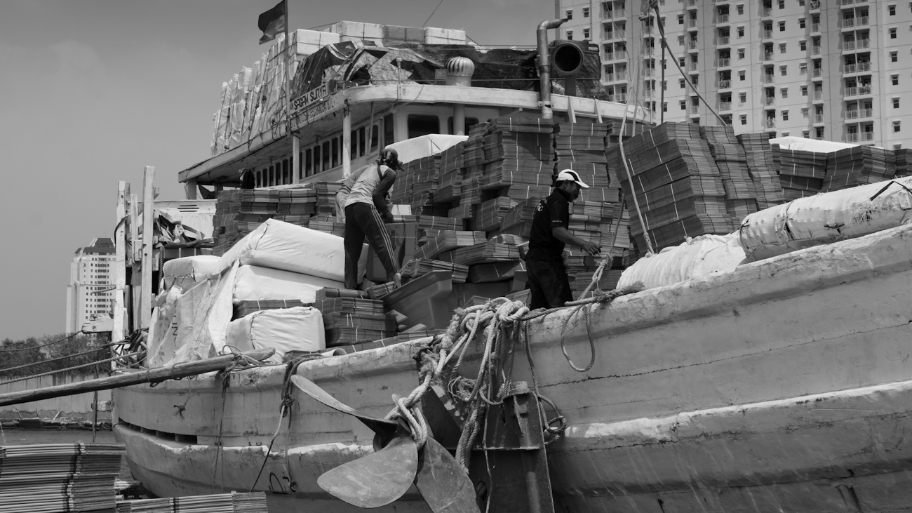 A pallet load of flat-packed cardboard boxes is loaded onto the deck of an awaiting Pinisi freighter using a deck crane.
