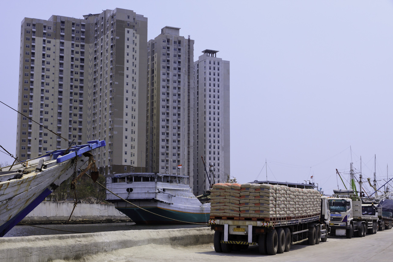 Freight trucks lined up on the wharf alongside motorised Pinisi at the old port of Sunda Kelapa, are towered over by apartment buildings.