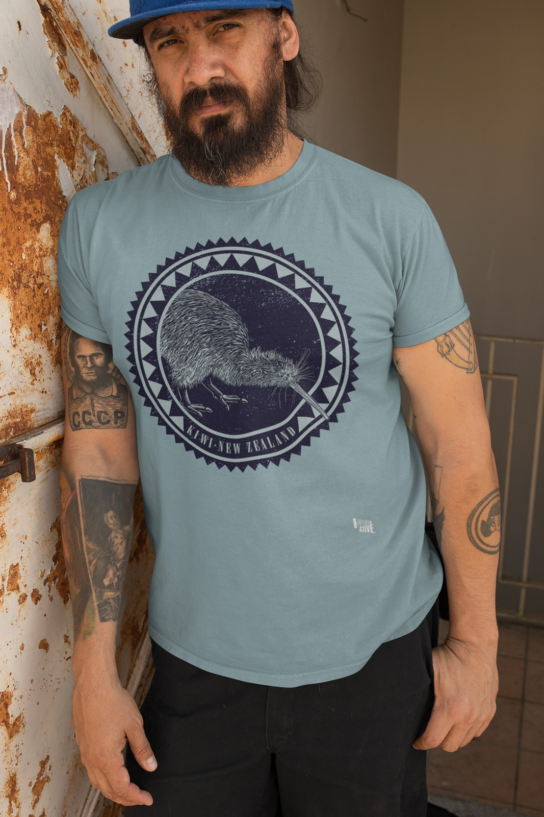 A bearded man wearing a heathered pacific grey kiwi New Zealand t-shirt leaning against a rusty wall.