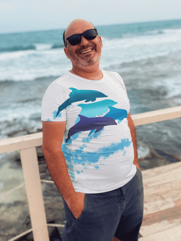 A man wearing a leaping dolphin all over print white teeshirt by the beach.