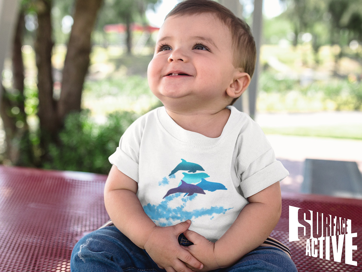 A baby boy looking up while smiling wearing a dolphins leaping New Zealand t-shirt.