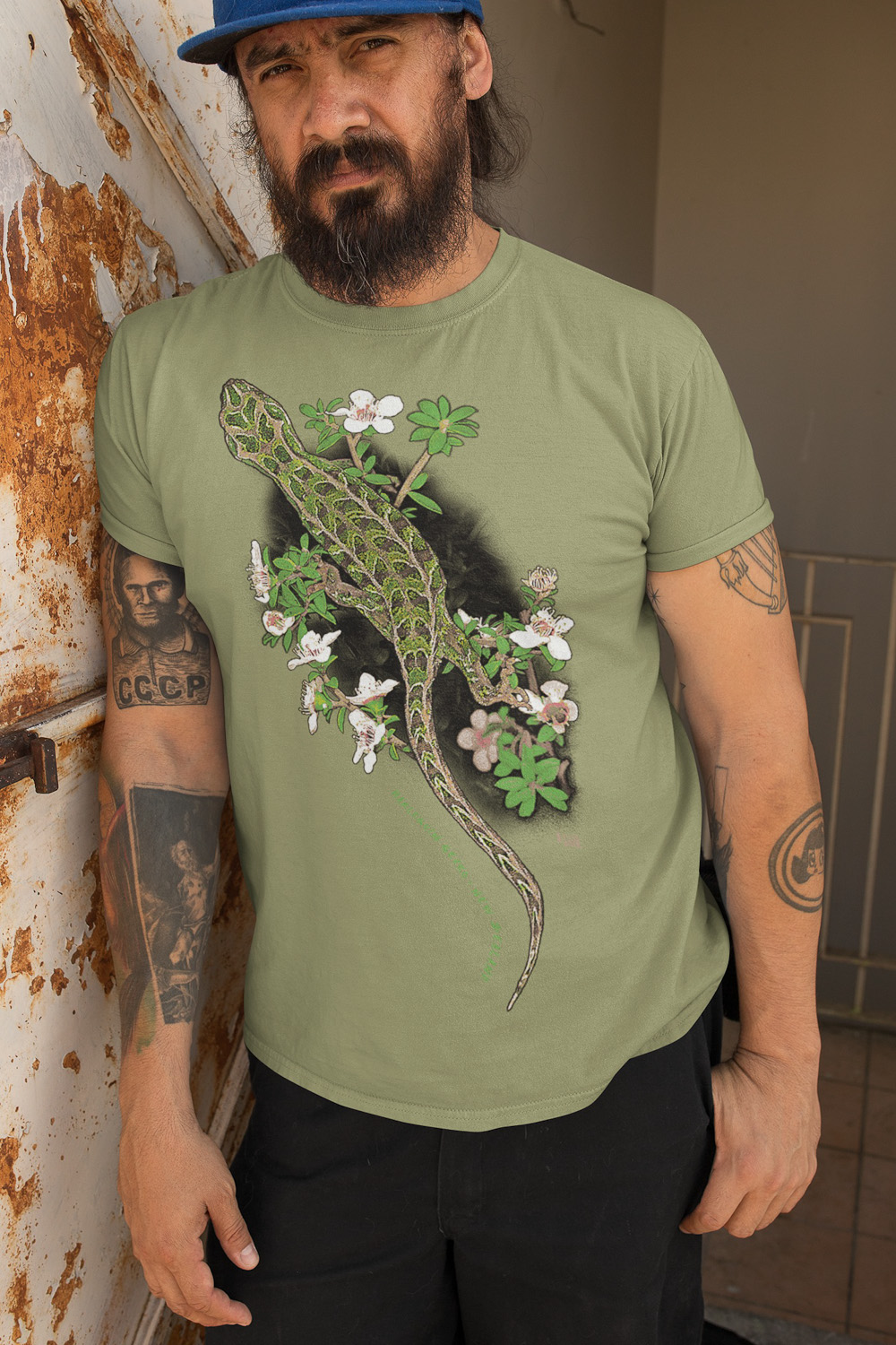 Close-up of a bearded man wearing a sage green t-shirt with a Harlequin Gecko design, leaning against a rusty wall.