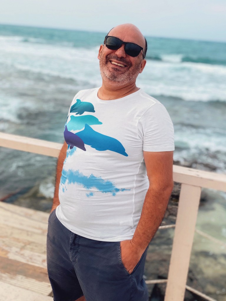 A happy middle aged man wearing a dolphins leaping New Zealand all-over printed t-shirt standing beside the sea.