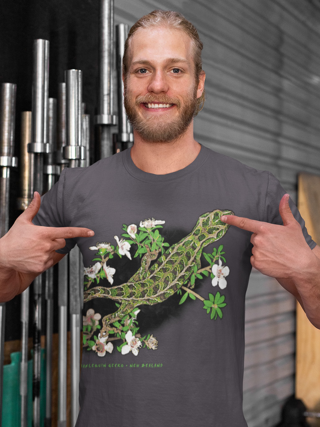 A man shows off a t-shirt with a Harlequin Gecko design, standing in front of gym weight lifting bars, showcasing the artful depiction of the gecko’s pattern.