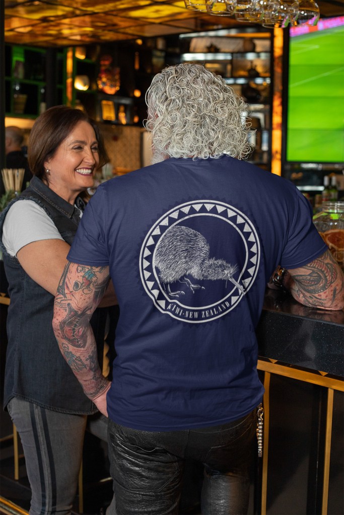 A white-haired man in a bar, wearing a navy t-shirt with a large, dark kiwi roundel print on the back.