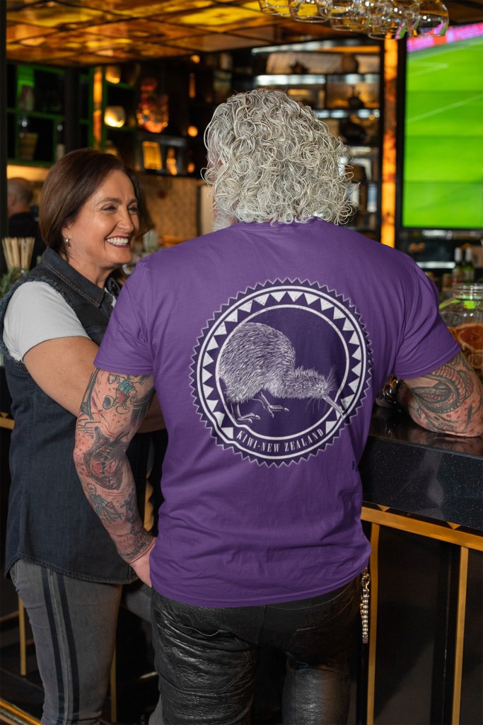 Back view of a white-haired man in a bar, wearing a violet t-shirt with a dark kiwi roundel print.