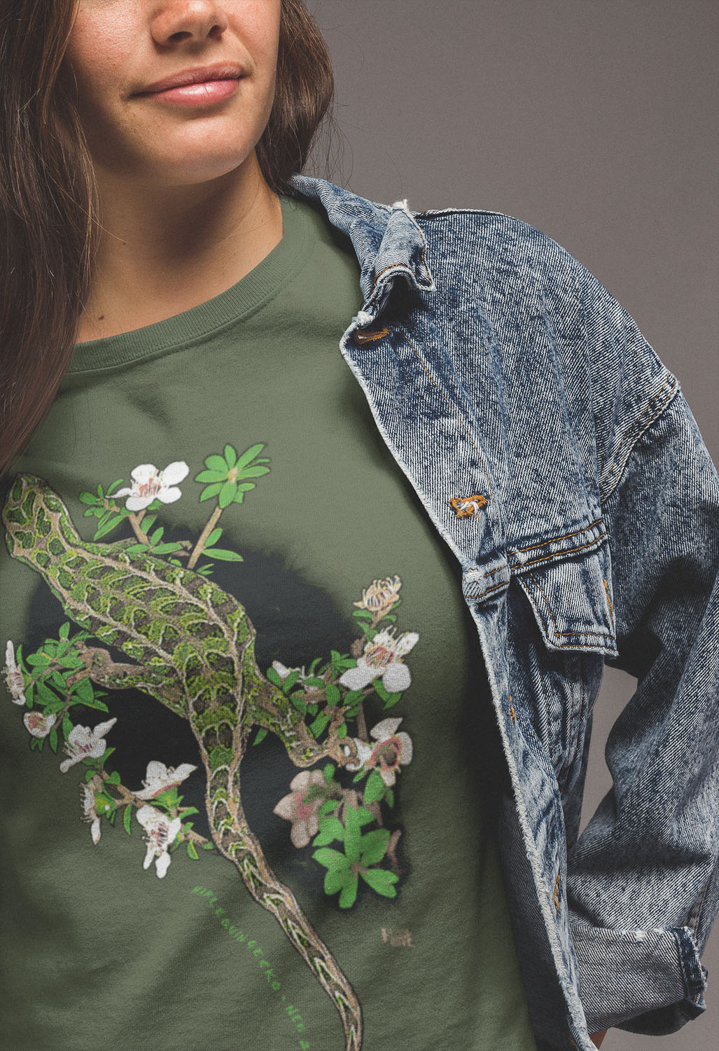 :Close-up of a woman wearing a sage green Harlequin Gecko t-shirt under a denim jacket, highlighting the detailed print of the gecko's camouflage pattern against a backdrop of flowering manuka.
