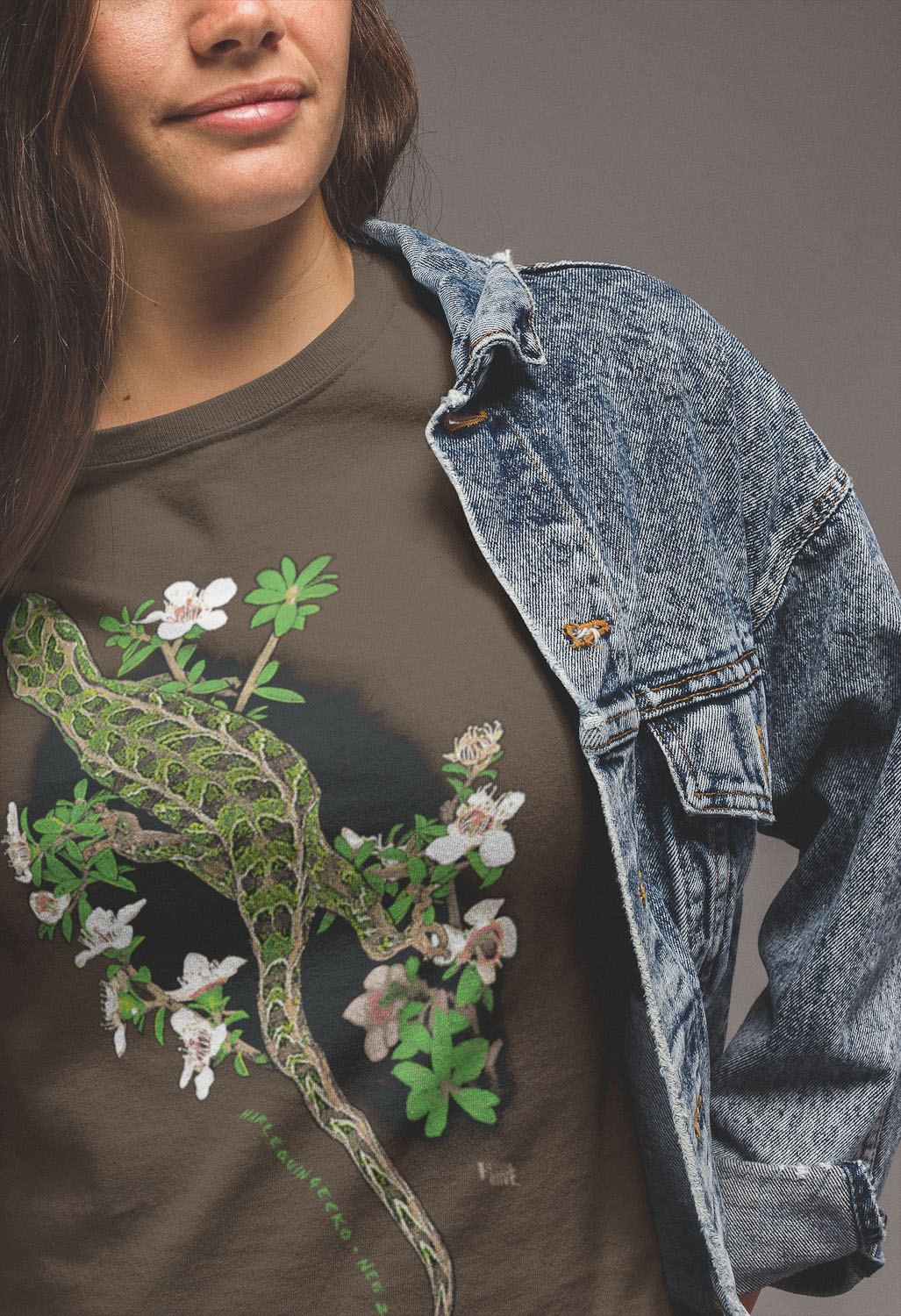 Close-up of a woman wearing a brown Harlequin Gecko t-shirt under a denim jacket, highlighting the detailed print of the gecko's camouflage pattern against a backdrop of flowering manuka.
