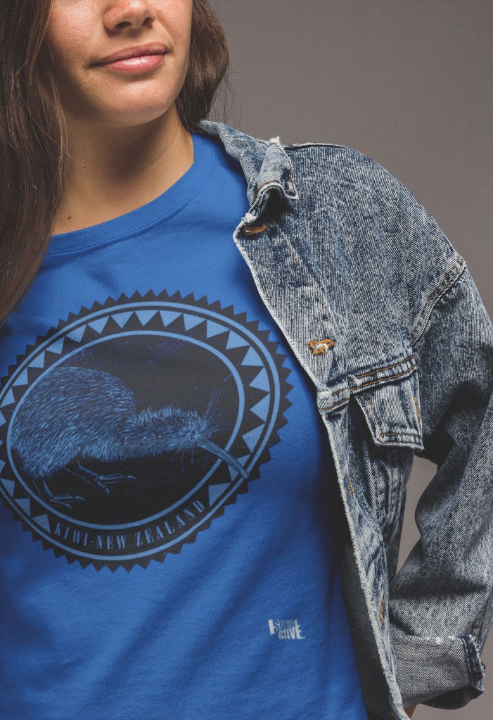 Close-up of a woman wearing a bright royal blue t-shirt with a dark Kiwi roundel print, layered under a denim jacket, in a studio setting.