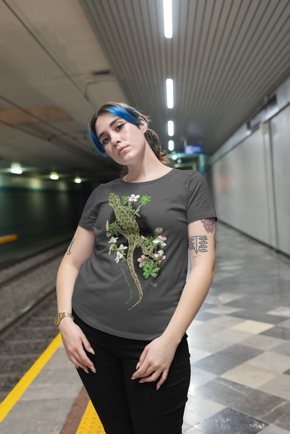 A plus-size woman with blue hair wears a Harlequin Gecko t-shirt featuring its distinctive pattern, standing in a subway station.
