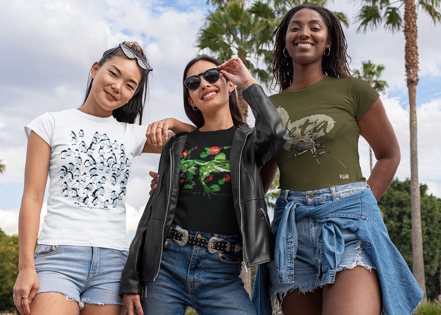 Three women smile brightly, each wearing a different Surface Active t-shirt showcasing New Zealand wildlife: an Adélie penguin, a jewelled gecko, and a giant weta.