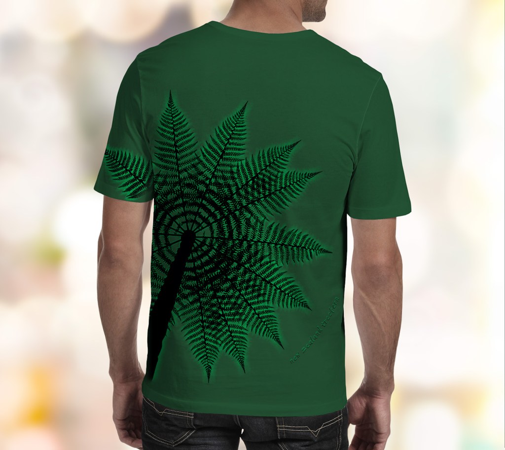 A forest green Surface Active all-over printed Treefern New Zealand t-shirt worn by a young man with his back to the camera in the studio.