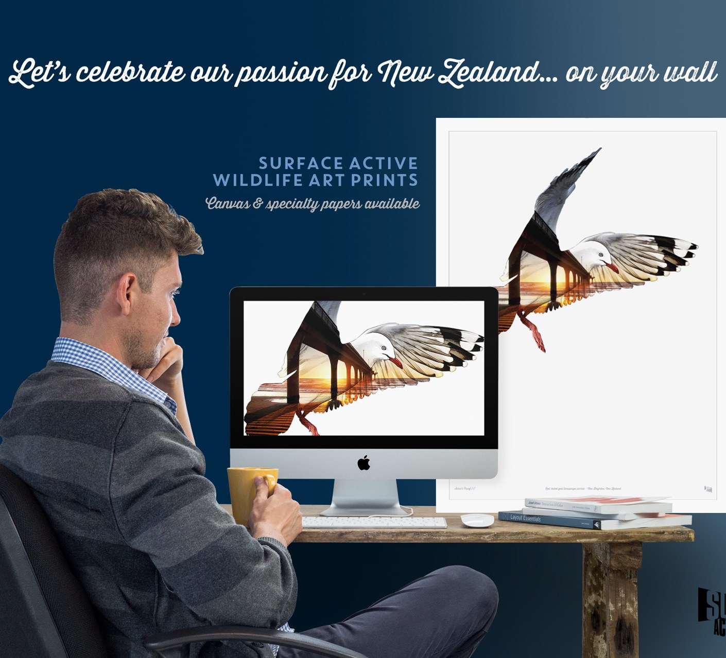 Photo of a young man with a cup of coffee engaged in art print production with a red-billed gull, New Brighton pier sunrise composite illustration on the computer monitor of his iMac alonside a matching large format framed poster of the same print sitting on his desk. The headline is on the dark navy blue background of the wall and reads: “Let's celebrate our passion for New Zealand… on your wall. Surface Active wildlife art prints. Canvas & specialty papers available.”