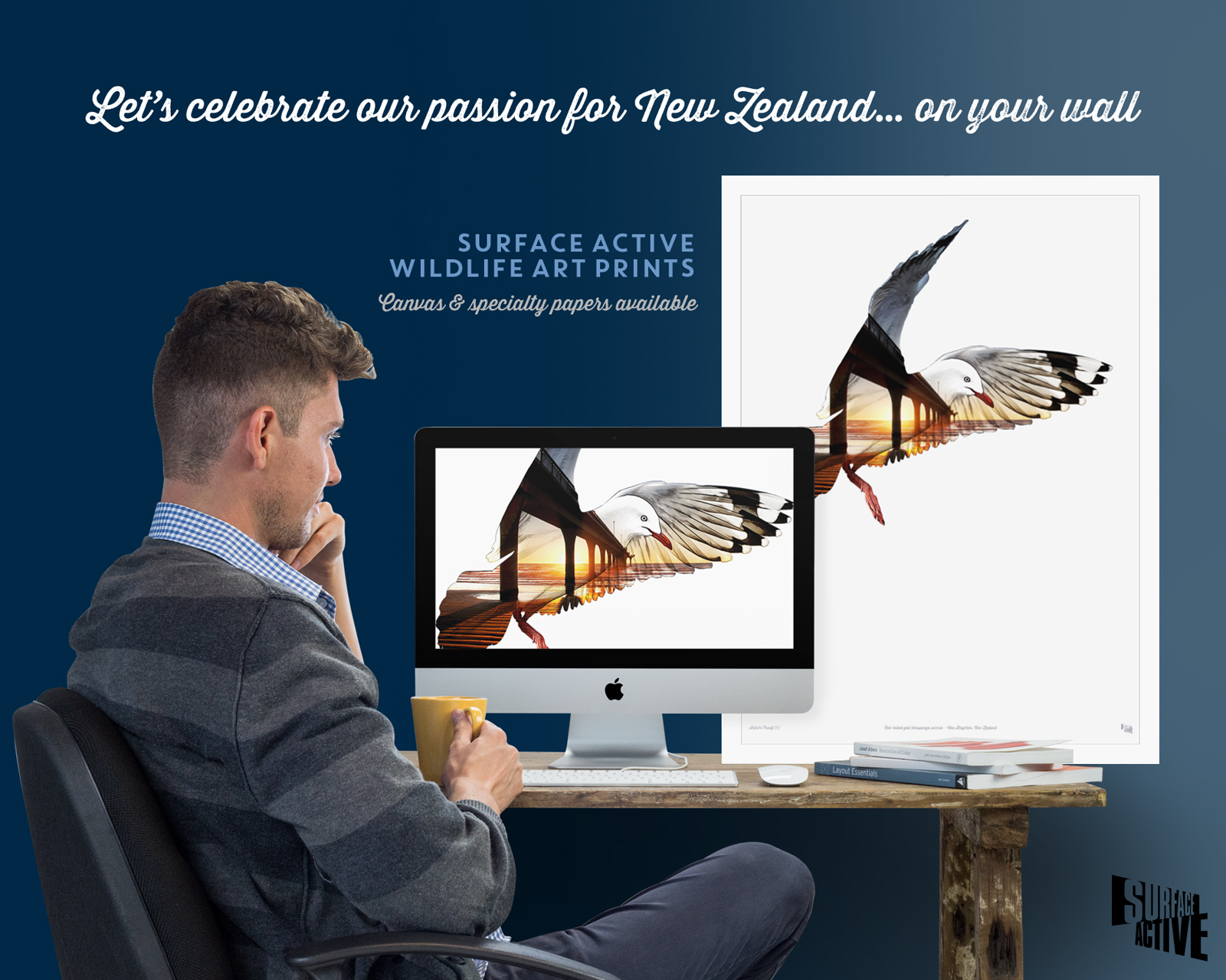 Photo of a young man with a cup of coffee engaged in art print production with a red-billed gull, New Brighton pier sunrise composite illustration on the computer monitor of his iMac alonside a matching large format framed poster of the same print sitting on his desk. The headline is on the dark navy blue background of the wall and reads: “Let's celebrate our passion for New Zealand… on your wall. Surface Active wildlife art prints. Canvas & specialty papers available.”