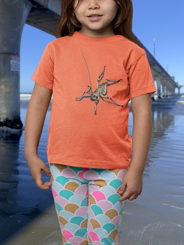 A joyful young girl wearing a bright orange t-shirt with an all-over weta print stands next to New Brighton Pier, with the ocean and sky behind her.