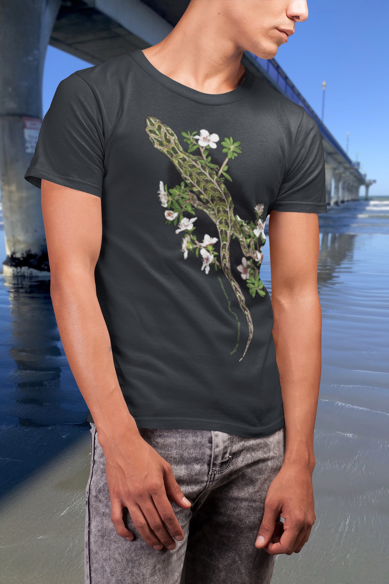 A multicolour print of an overhead view of a Harlequin Gecko moving through the foliage of a flowering manuka tree printed on the front of a black marle Surface Active t-shirt. The shirt reads in small text “Harlequin gecko - Stewart Island New Zealand.