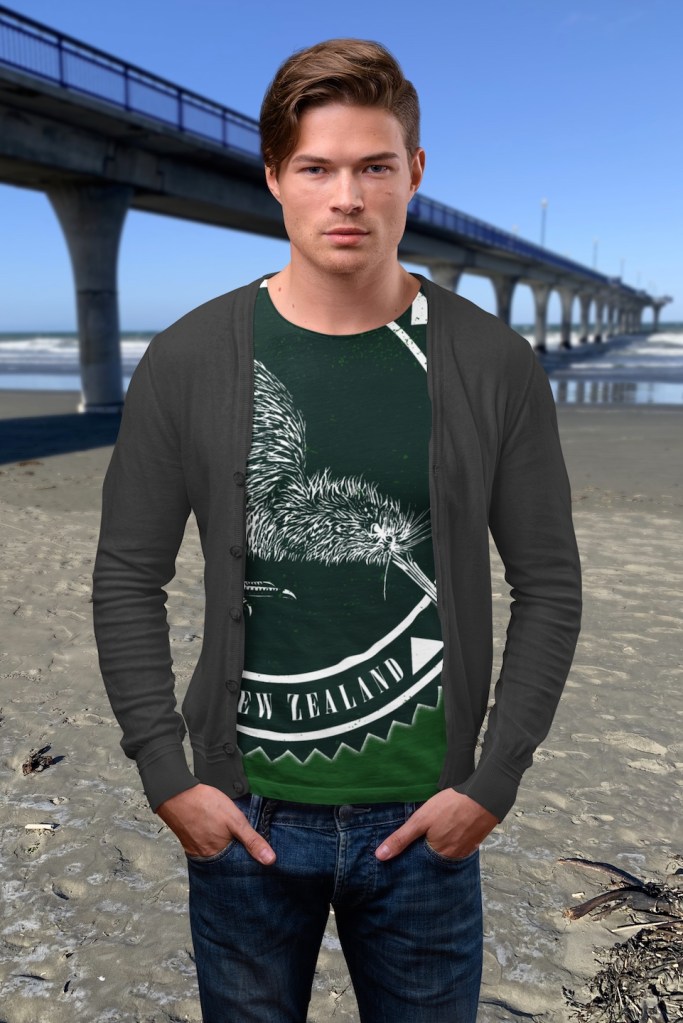 A man wearing a Surface Active all-over printed iconic Kiwi, New Zealand t-shirt, dark coloured shirt version, standing on New Brighton beach beside the pier.