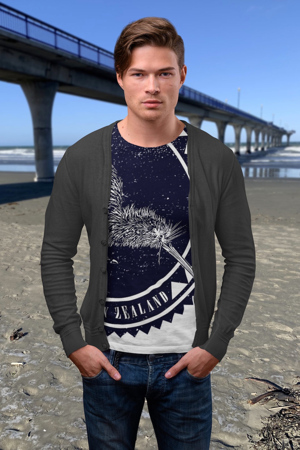 A man wearing a Surface Active all-over printed iconic Kiwi, New Zealand t-shirt, light coloured shirt version, standing on New Brighton beach beside the pier.